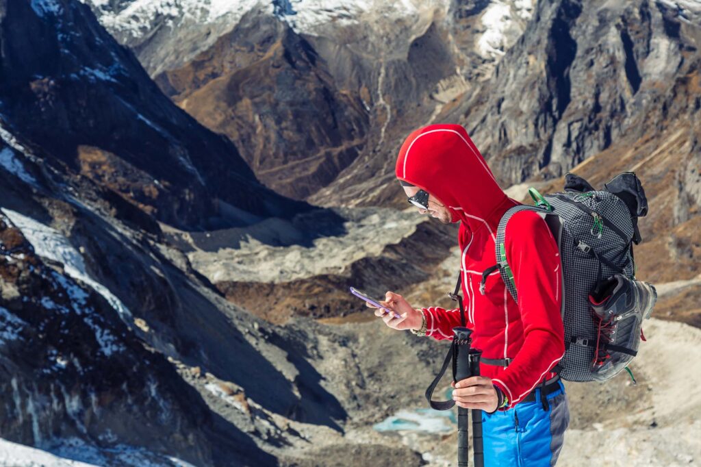 Bergsteiger in roter Jacke steht auf einem Gipfel und sieht auf sein Smartphone