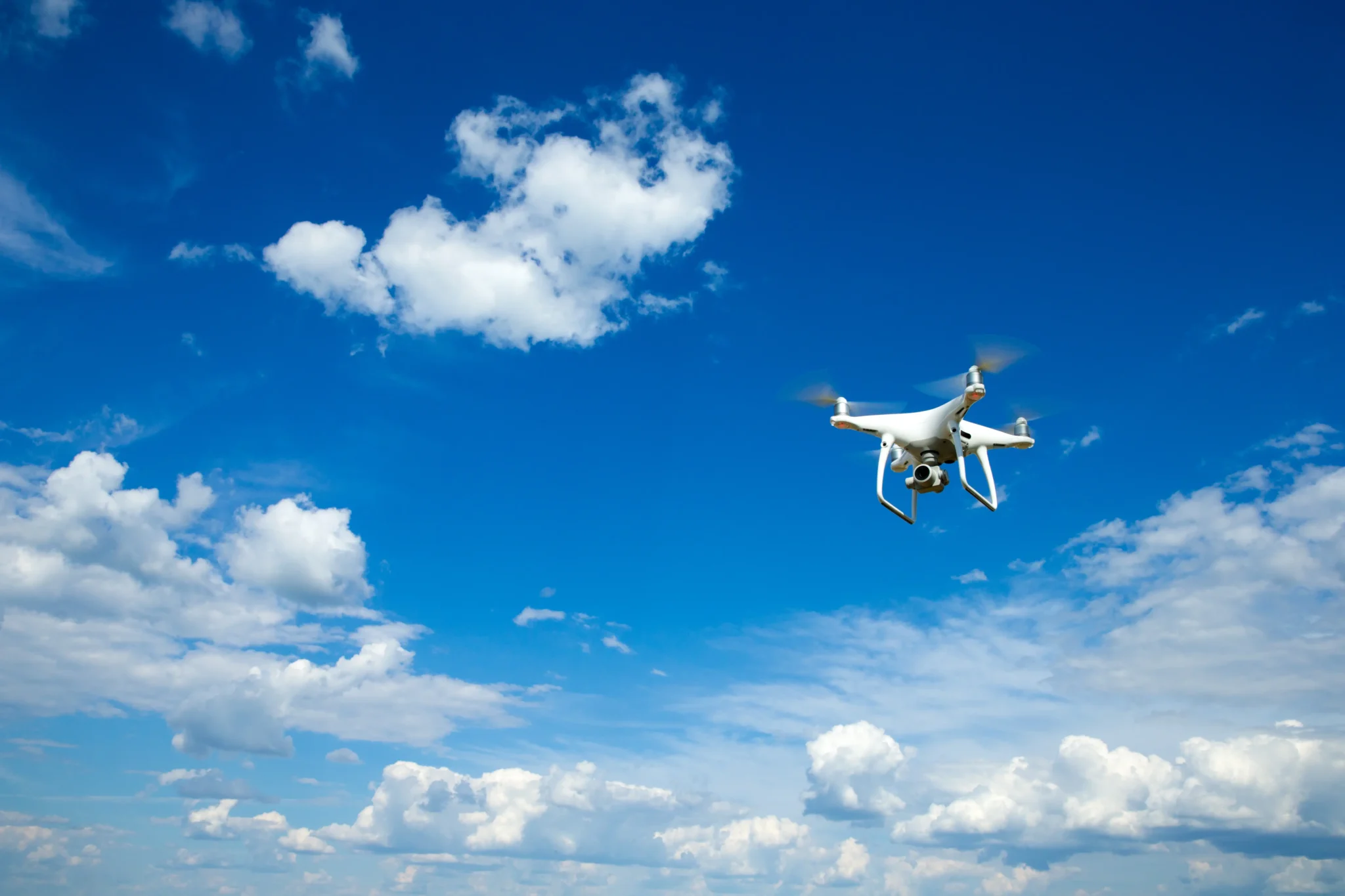 Weiße Kameradrohne fliegt bei klarem, blauem Himmel mit vereinzelten Wolken – Symbol für moderne zivile Drohnentechnologie und mobile Kommunikation aus der Luft.
